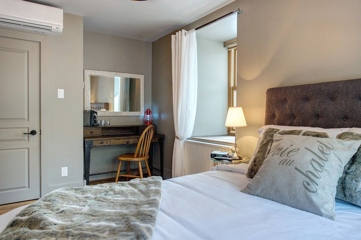 Une chambre à coucher comprenant un lit, un bureau, une chaise, un miroir, un radiateur et une fenêtre avec des rideaux.
