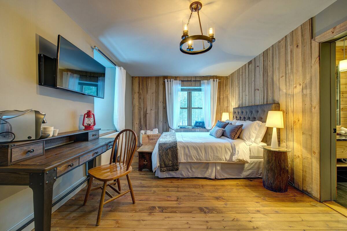 Une chambre avec un plancher en bois, un lit avec une literie blanche, un bureau avec une chaise, un téléviseur fixé au mur et une fenêtre avec des rideaux.