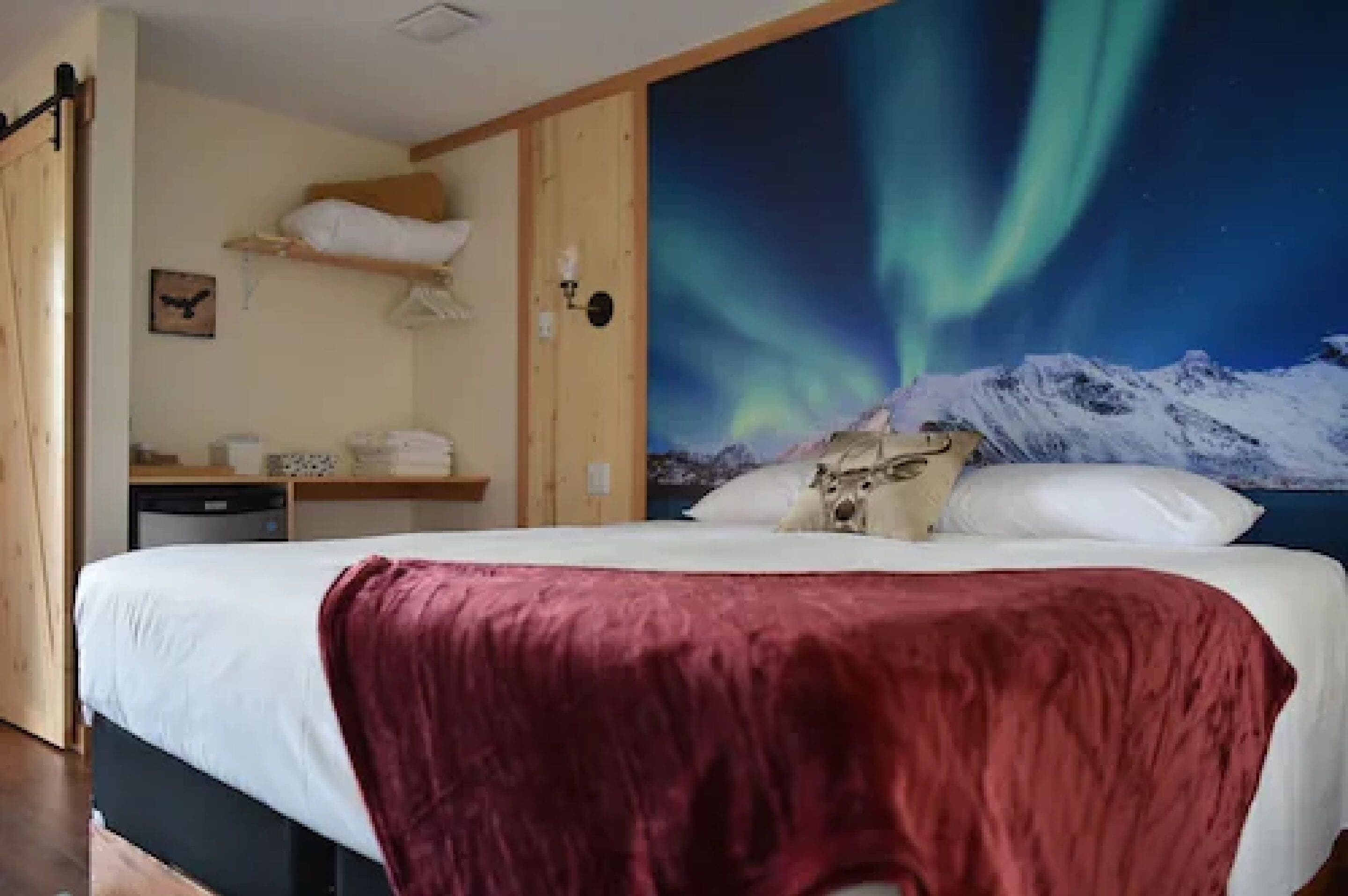 Un dormitorio con cama, mesita de noche, estante en la pared y un cuadro de las auroras boreales.