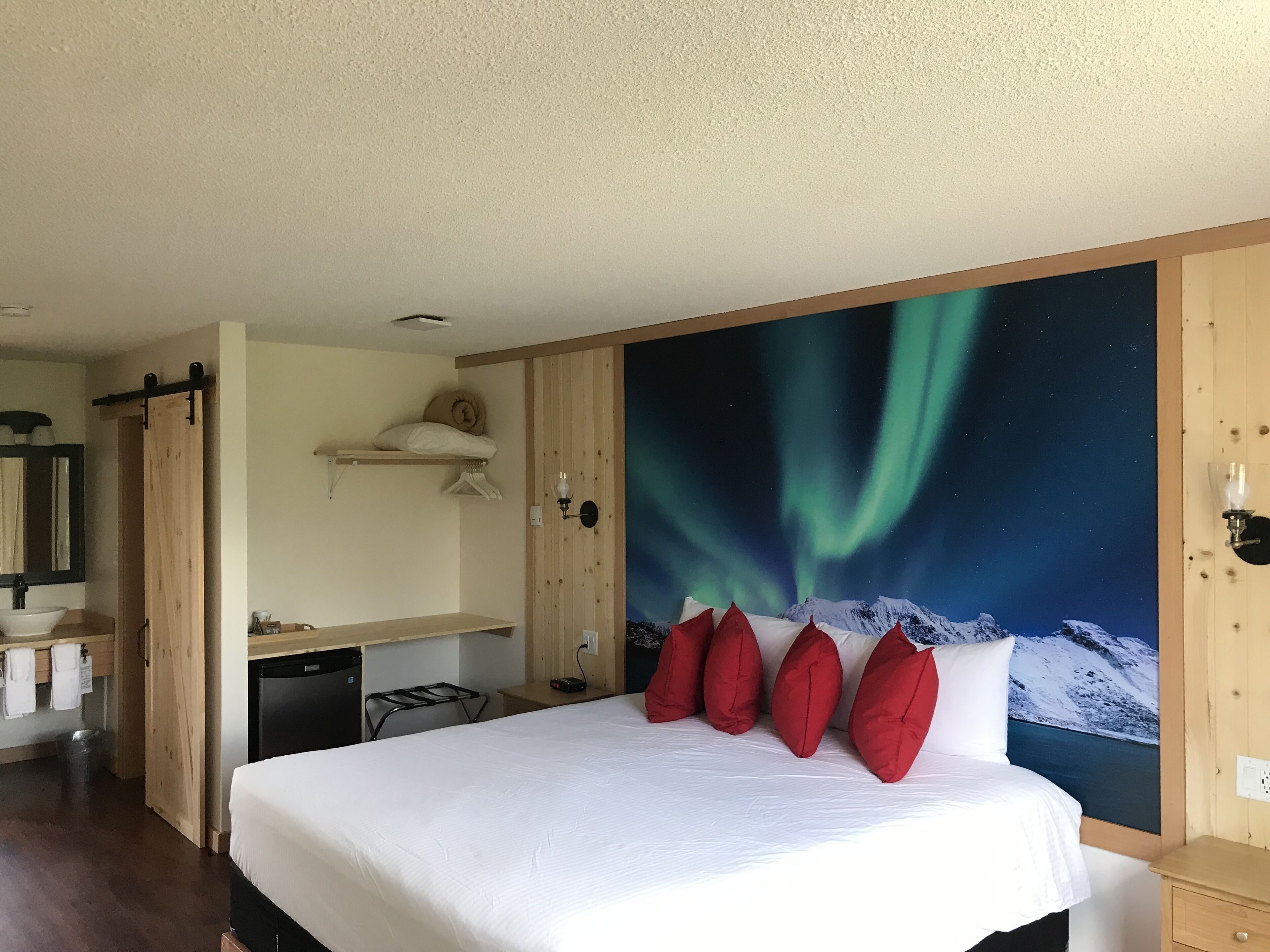 Habitación de hotel con cama, mesita de noche, estante de madera, espejo y un mural en la pared con la aurora boreal.