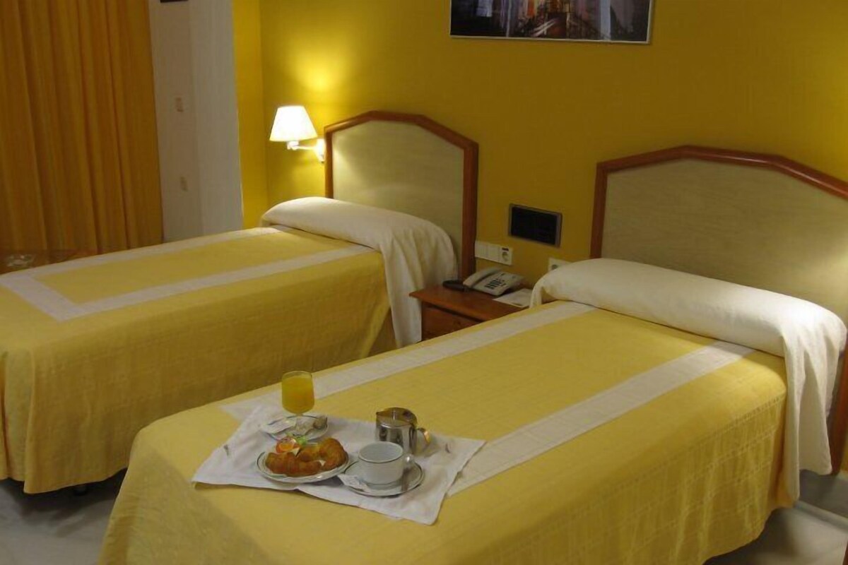 A hotel room with two single beds, each with a yellow bedspread and white pillows. There is a small table with a plate of croissants, a cup, and a glass of orange juice. A lamp is on the nightstand next to the beds.