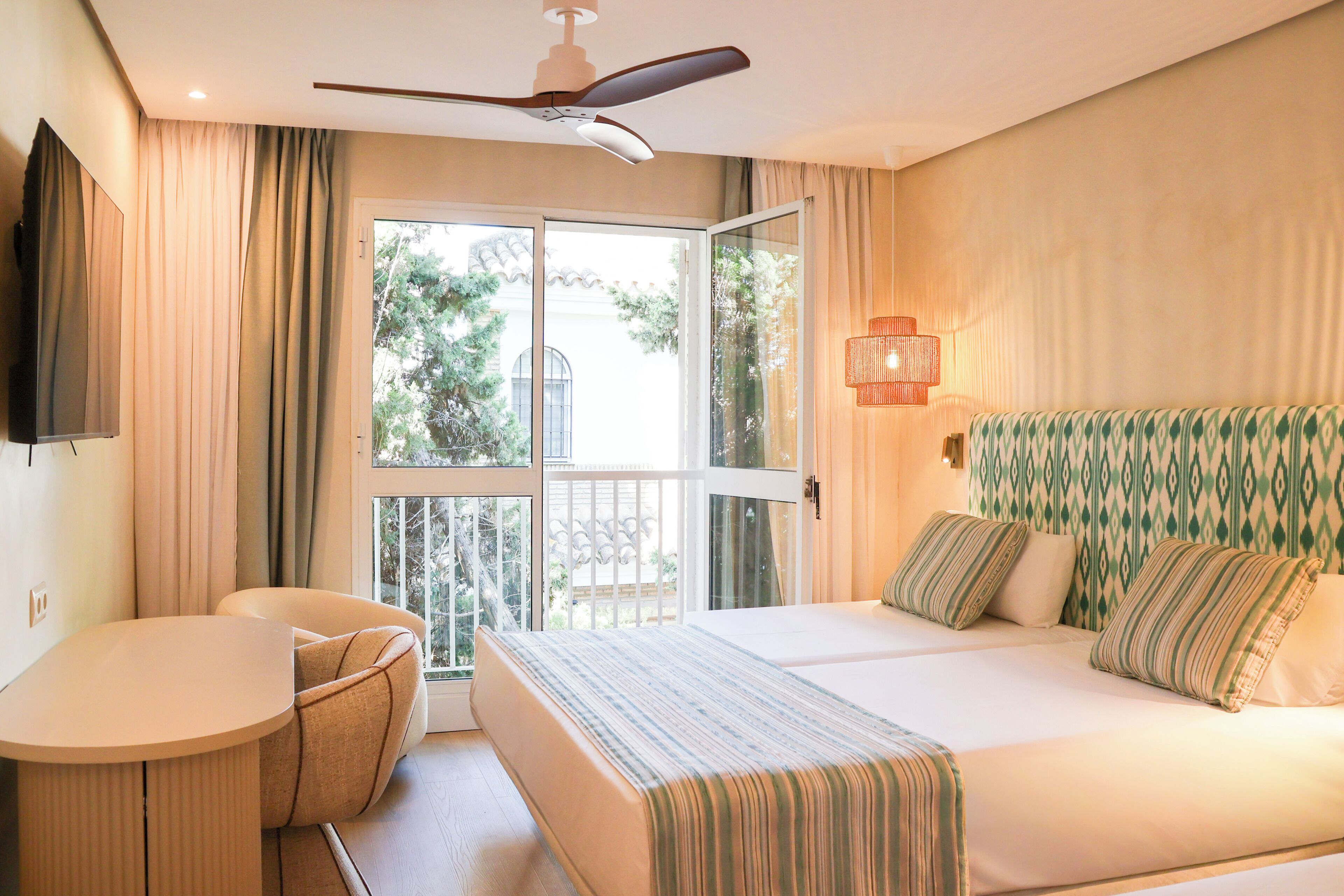 Habitación de hotel con cama, escritorio, silla, televisor y balcón con cortinas.