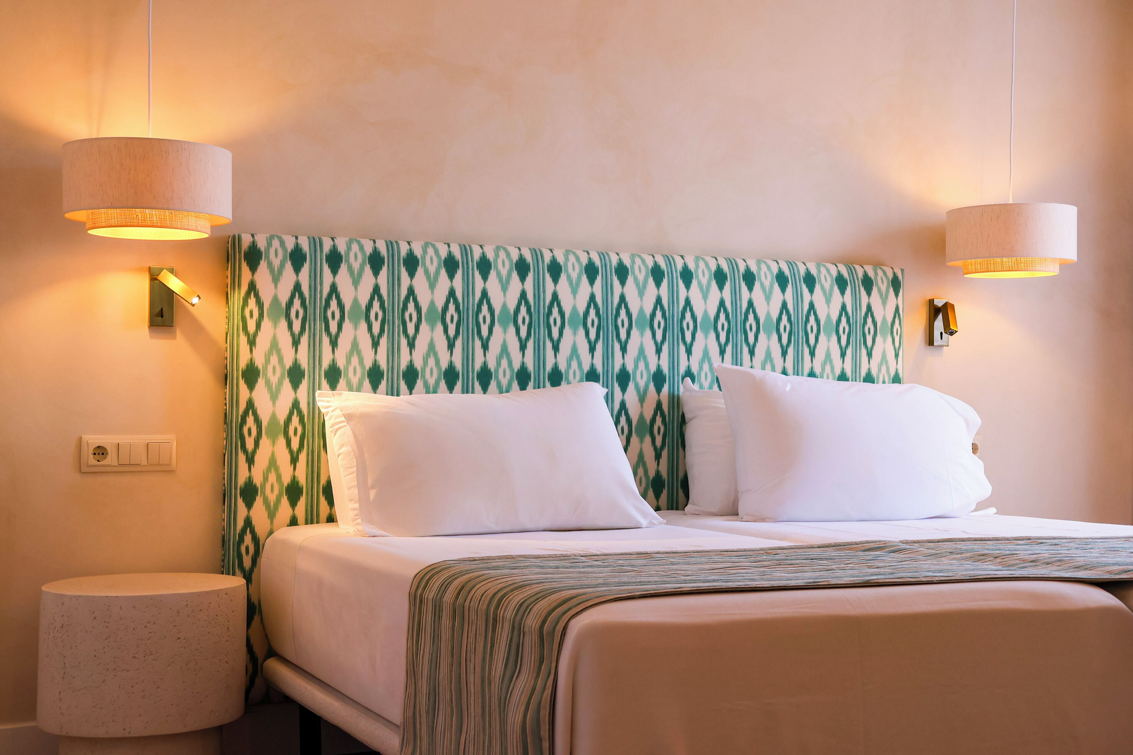 A hotel room with a bed, two wall-mounted lamps, a power outlet, and a textured wall.