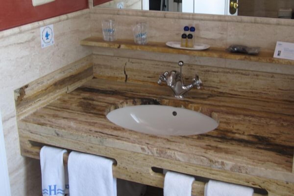 A bathroom with a wooden vanity, a white sink, and a mirror. There are towels hanging on the wall and a small shelf with toiletries.