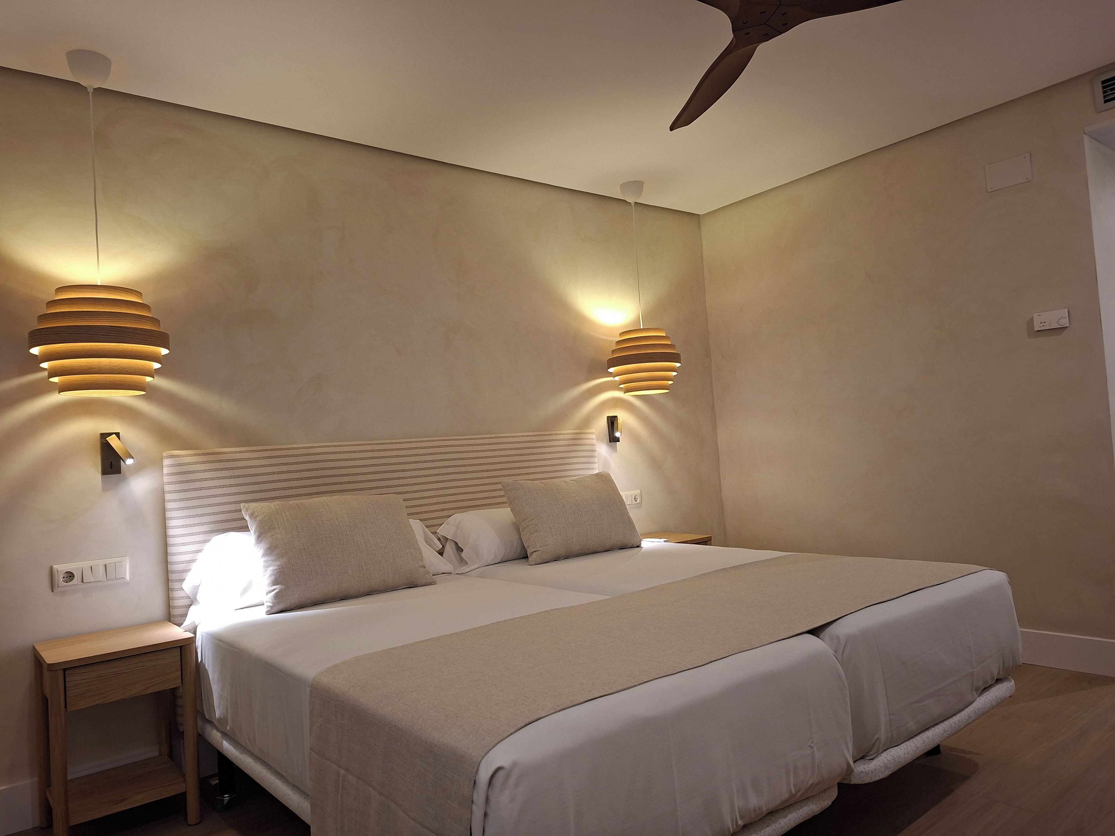 Une chambre à coucher avec un grand lit, deux tables de chevet, un ventilateur de plafond et des luminaires fixés au mur.