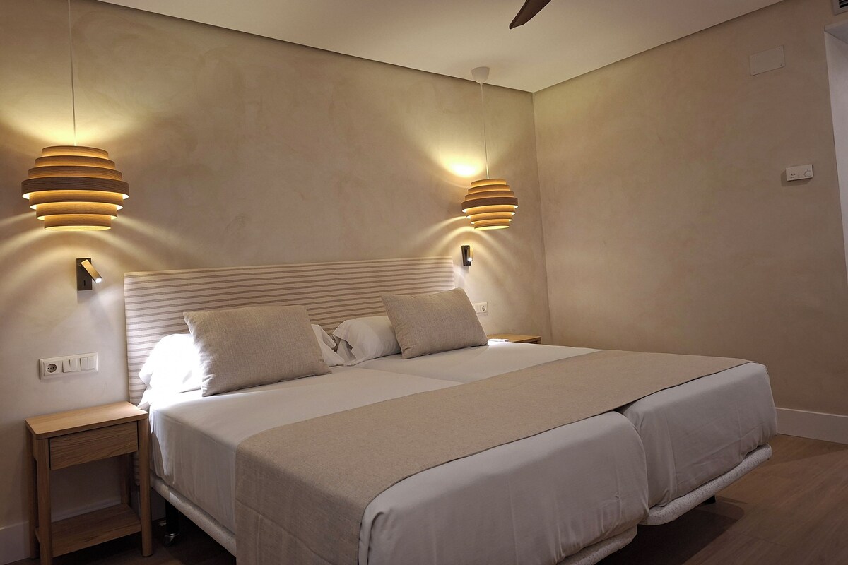 Un dormitorio con una cama grande, dos mesitas de noche, un ventilador de techo e iluminación en la pared.
