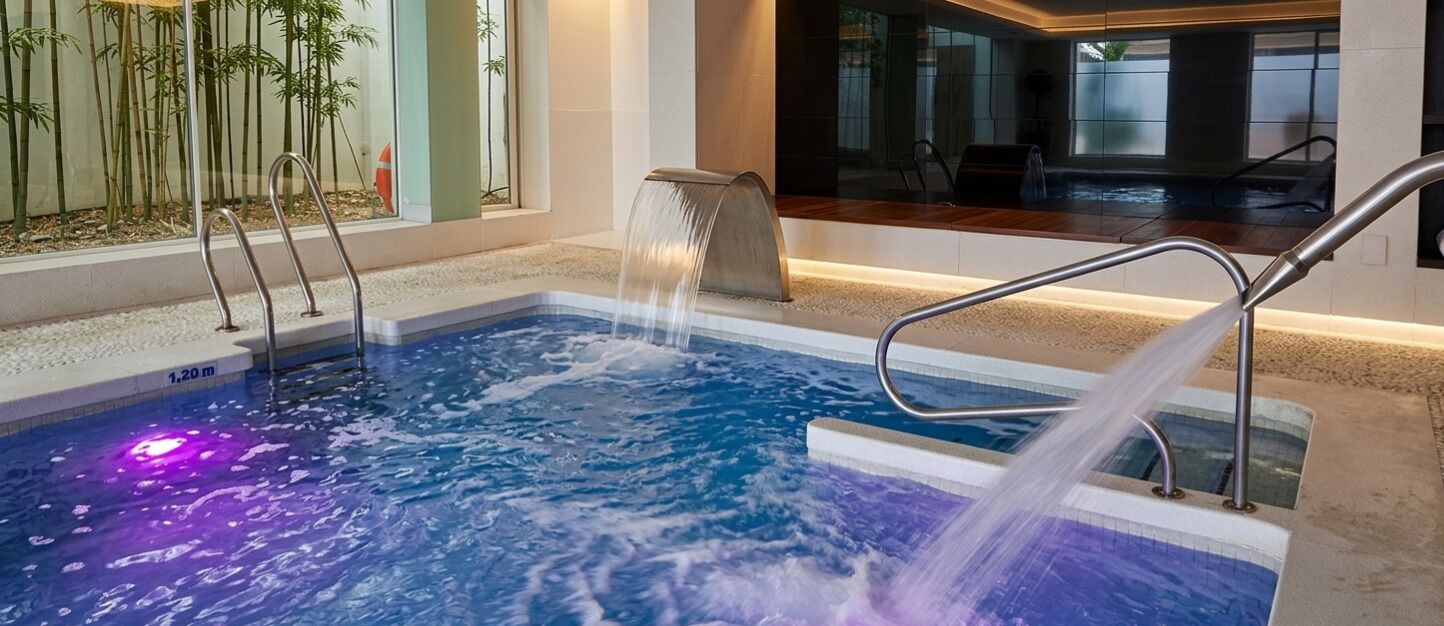 An indoor swimming pool with a waterfall feature, surrounded by a tiled area and a glass wall with bamboo plants.