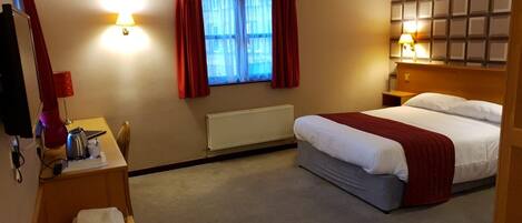 Habitación de hotel con cama, escritorio con lámpara, radiador y ventana con cortinas.