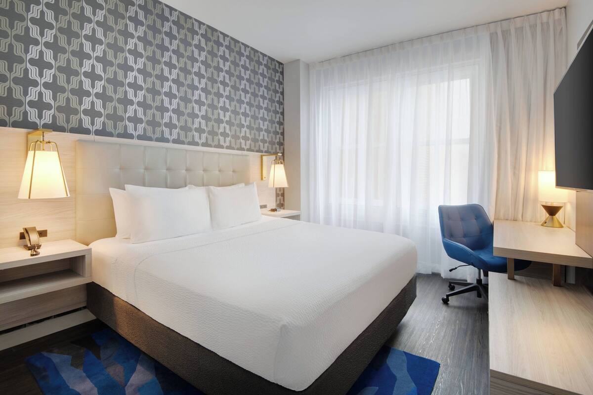 A hotel room with a large bed, two bedside tables with lamps, a desk with a chair, and a patterned wallpaper.