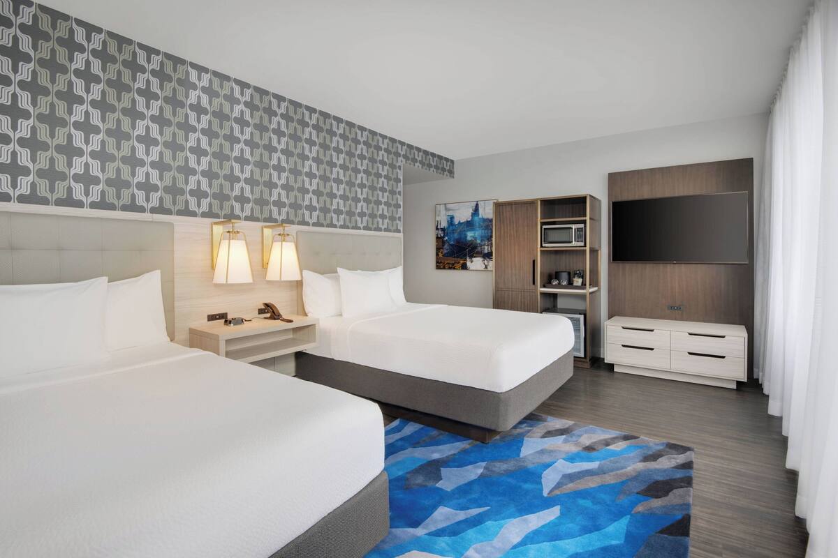 A hotel room with two beds, a desk with a lamp, a TV, a wardrobe, and a patterned wall.