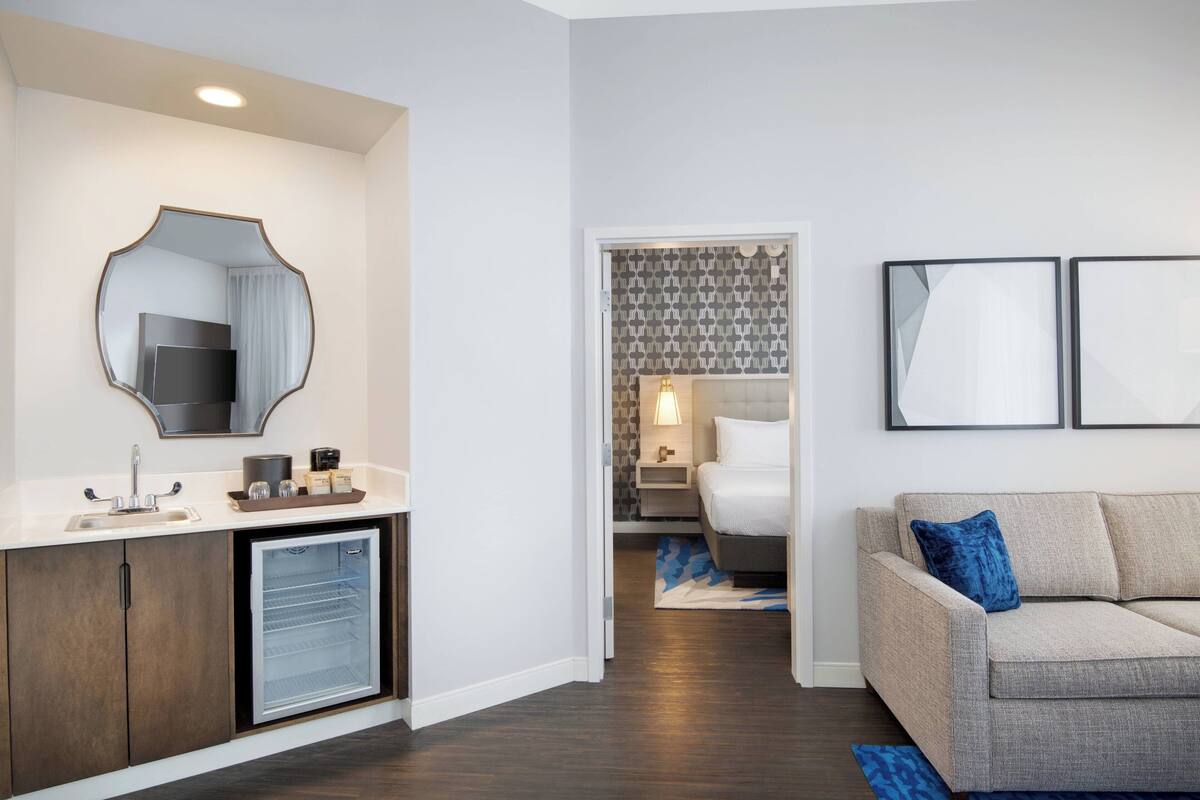 A modern hotel room with a kitchenette, a bed, a sofa, and a bathroom with a mirror and sink.