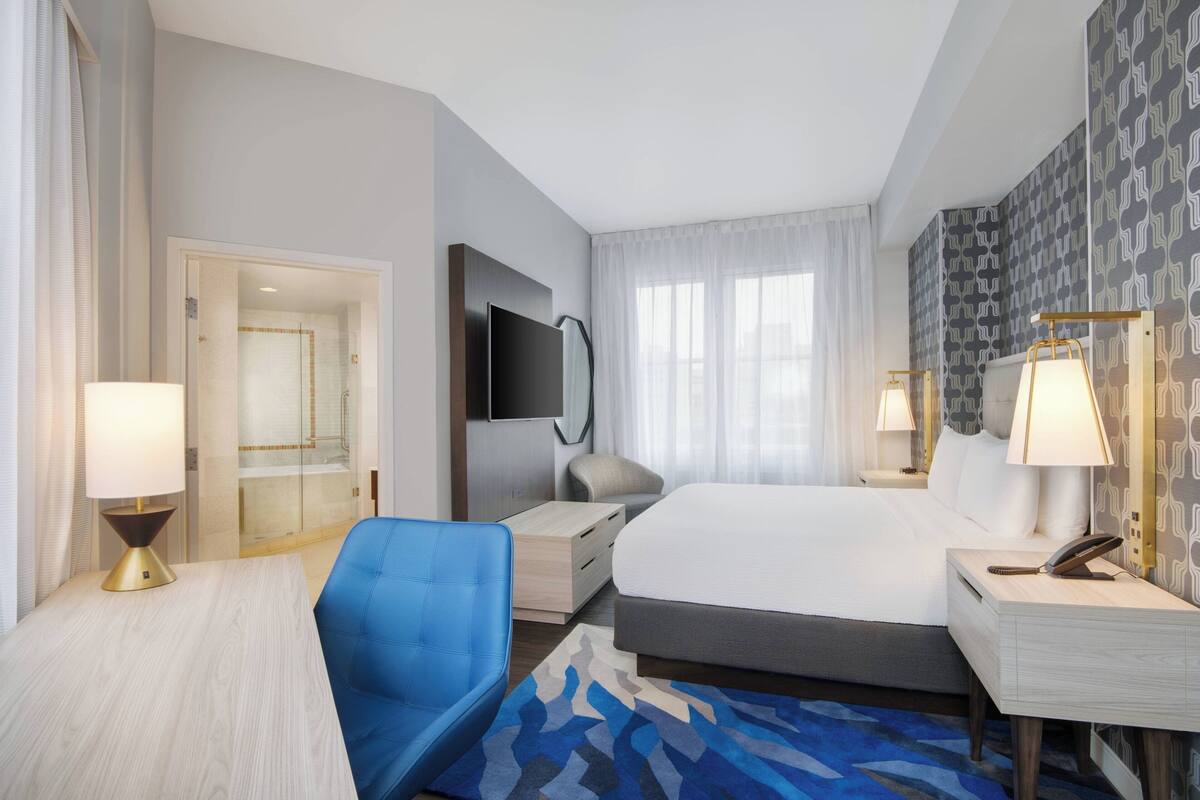 A hotel room with a bed, a desk, a chair, a TV, and a bathroom visible through an open door.