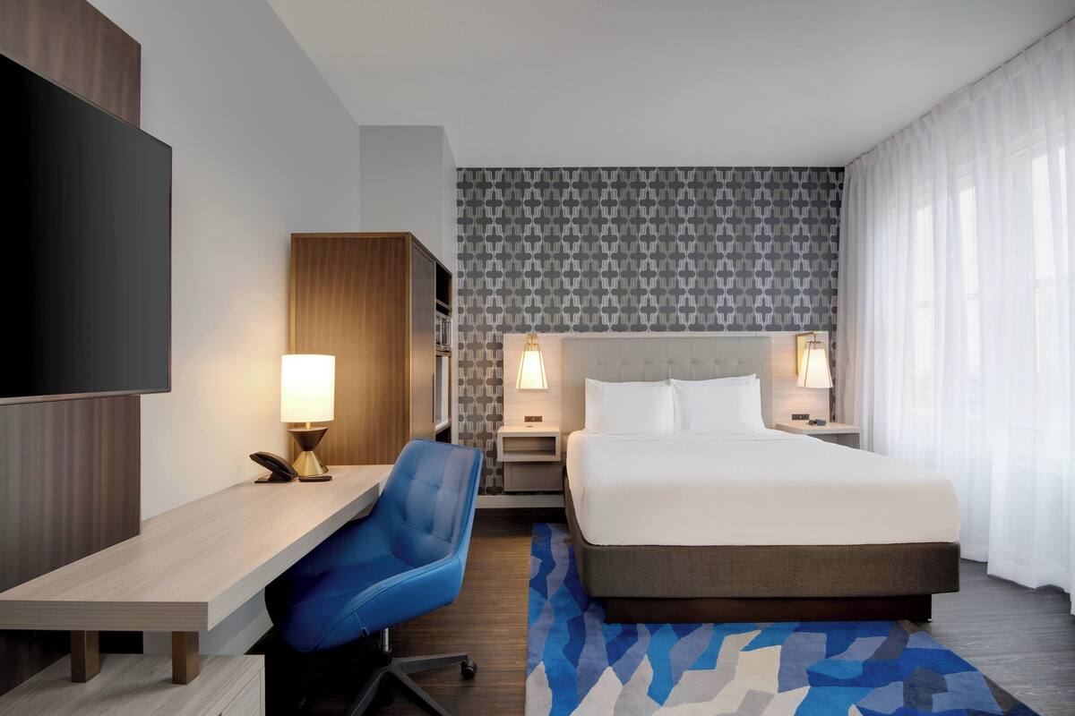 A hotel room with a bed, desk, chair, and a TV.