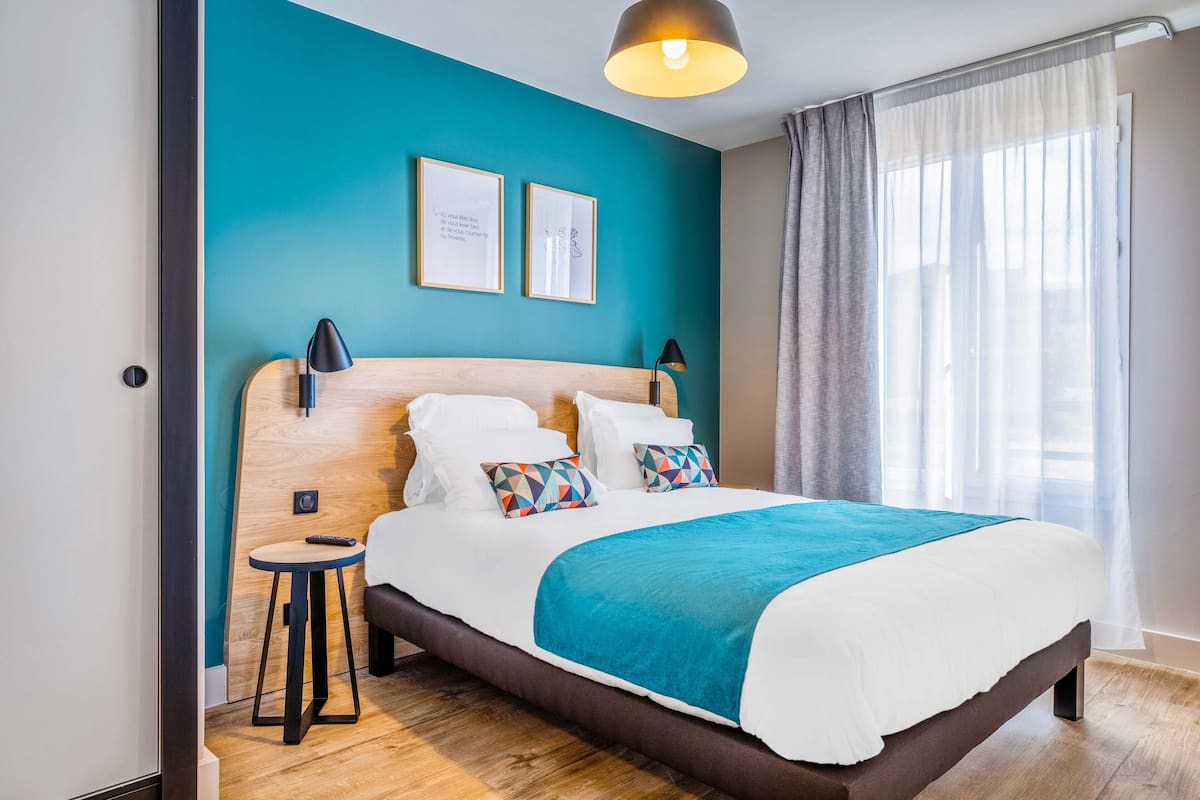 Ein modernes Hotelzimmer mit einem großen Bett, einem kleinen Beistelltisch und Blick ins Freie durch transparente Vorhänge.
