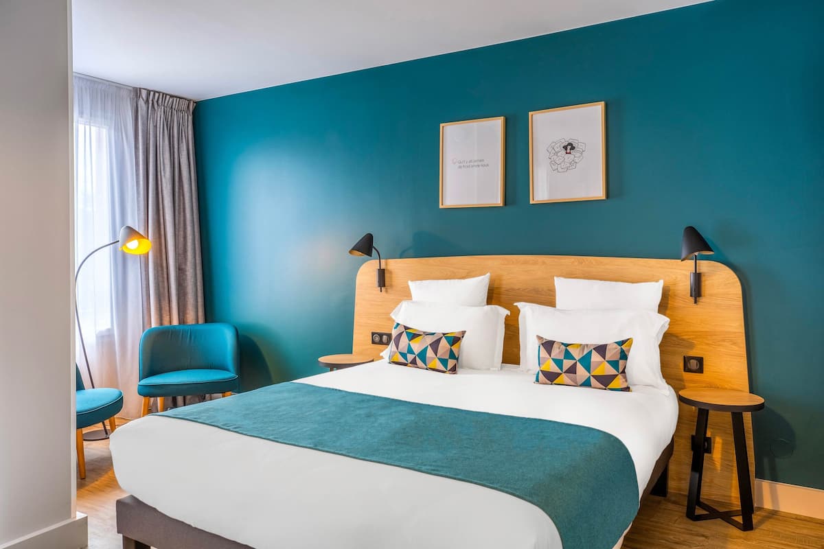 Ein modernes Hotelzimmer mit einer türkisfarbenen Akzentwand, einem großen Bett mit weißer Bettwäsche, zwei gerahmten Kunstwerken, einem blauen Sessel und einem Beistelltisch mit Lampe.