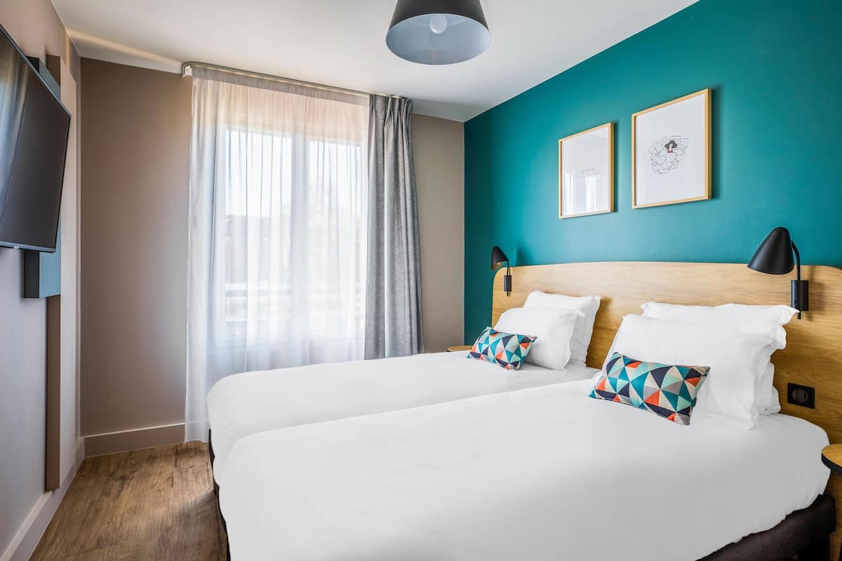 Ein Hotelzimmer mit zwei Betten, einem Fernseher, einem Fenster mit Vorhängen und zwei gerahmten Bildern an der türkisblauen Wand.
