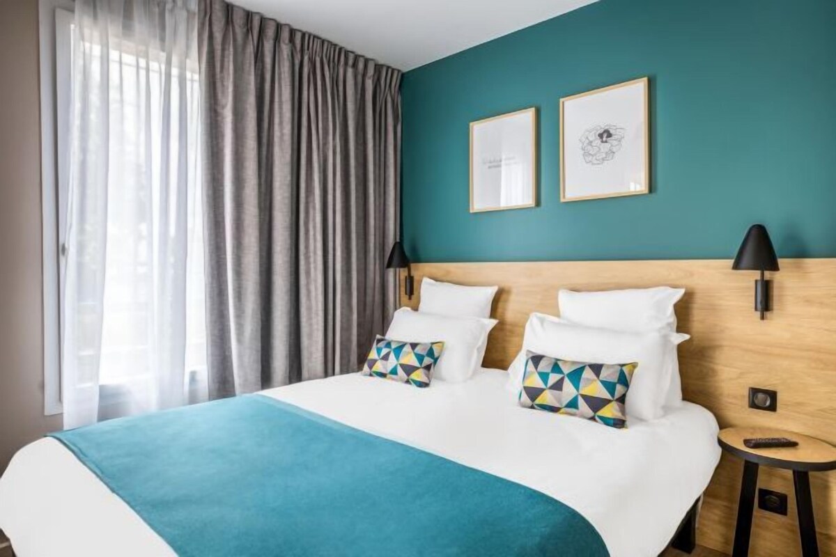 A hotel room with a bed, two wall-mounted lamps, a side table, and a window with curtains.