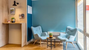 A modern interior with a blue accent wall, two light blue armchairs, a small round table with a plant, a wooden reception desk, and a ceiling with recessed lighting. - Appart'City Confort St Quentin en Yvelines - Bois d'Arcy (Bois-d'Arcy)