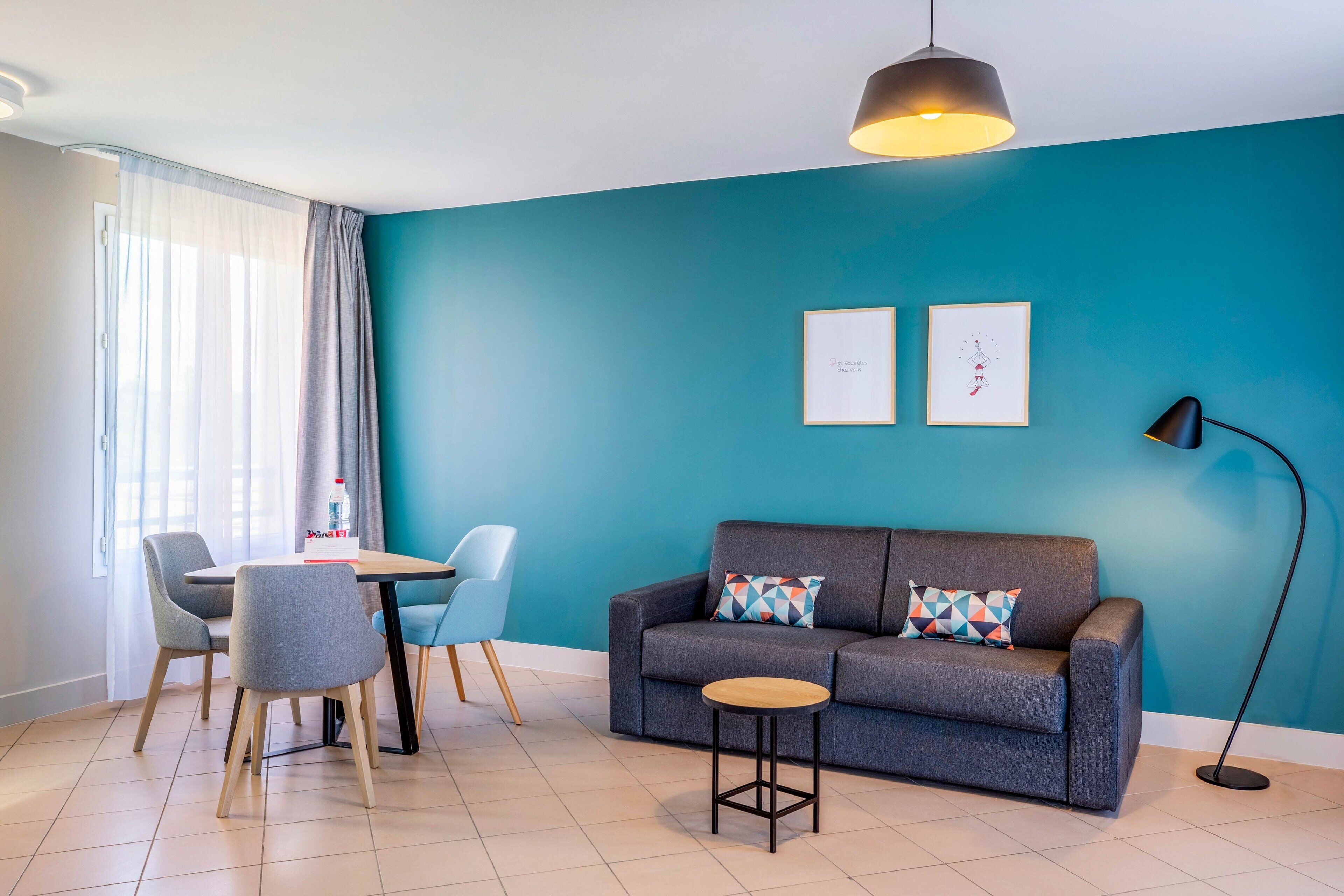 A modern living room with a teal accent wall, a grey sofa with patterned cushions, a round wooden side table, and a dining area with a white table and grey chairs.