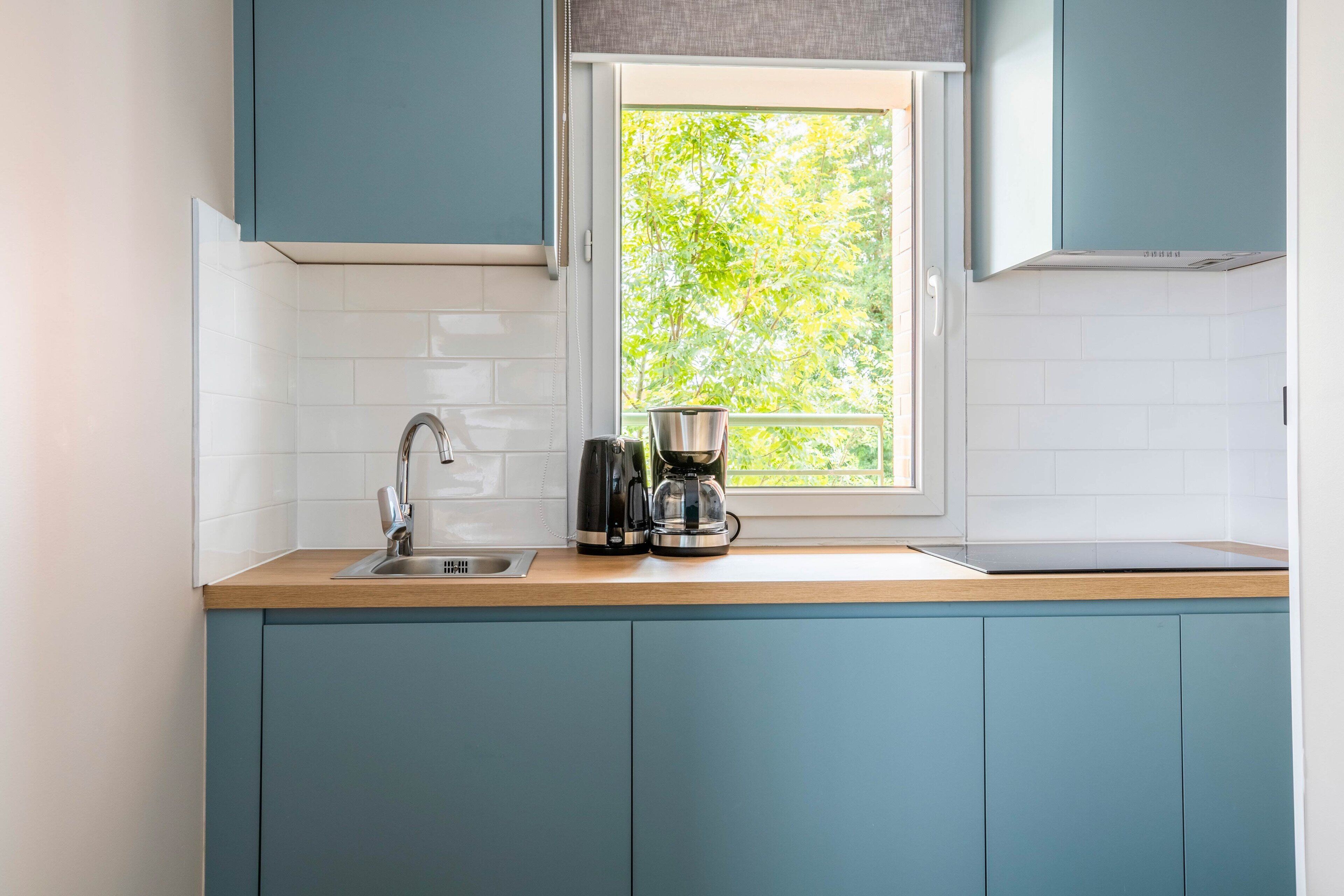 A kitchen with teal cabinets, a window with a view of greenery, a sink, and a coffee maker.