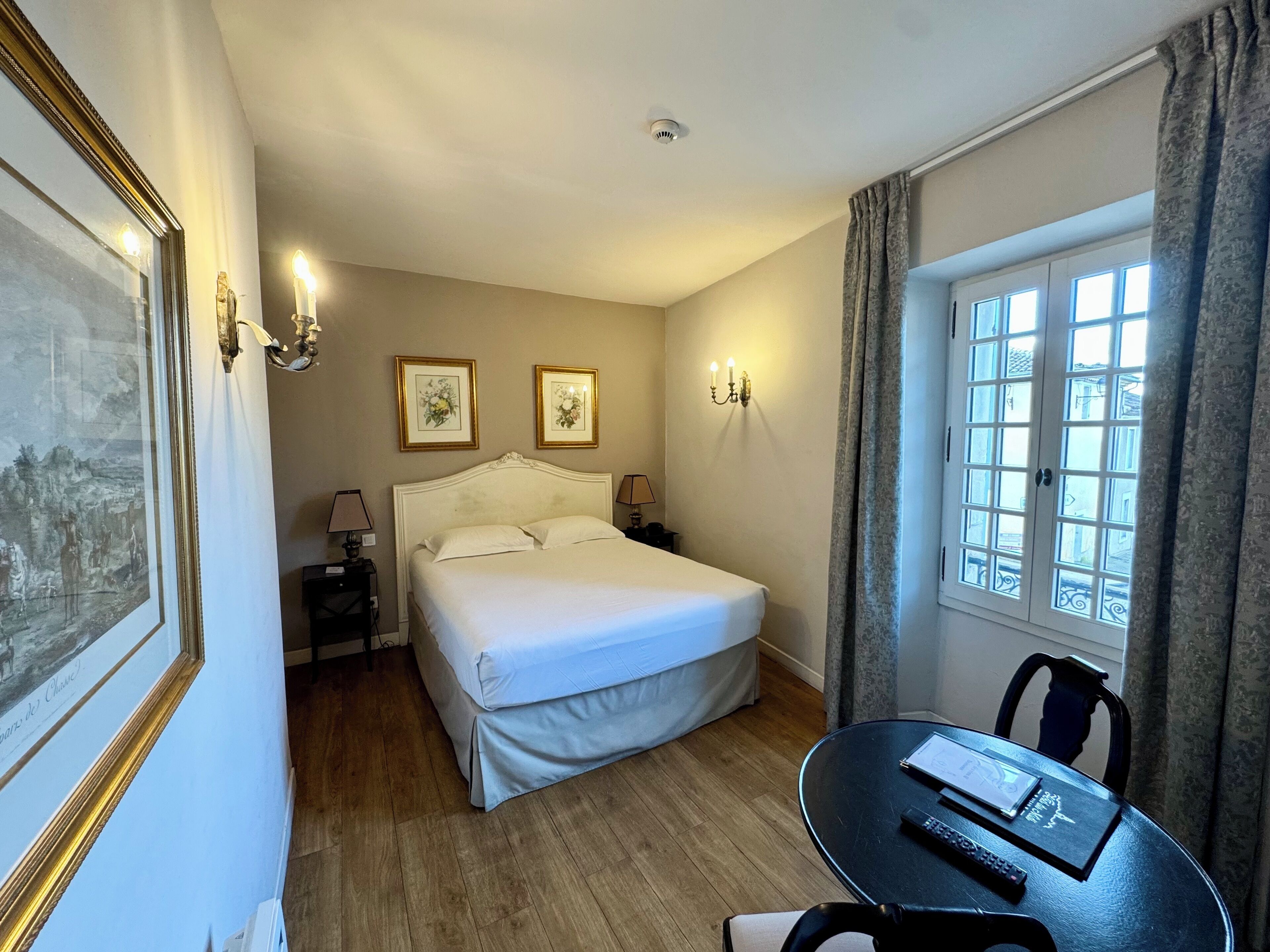 A hotel room with a large bed, a small table with a remote control, and a window with curtains.