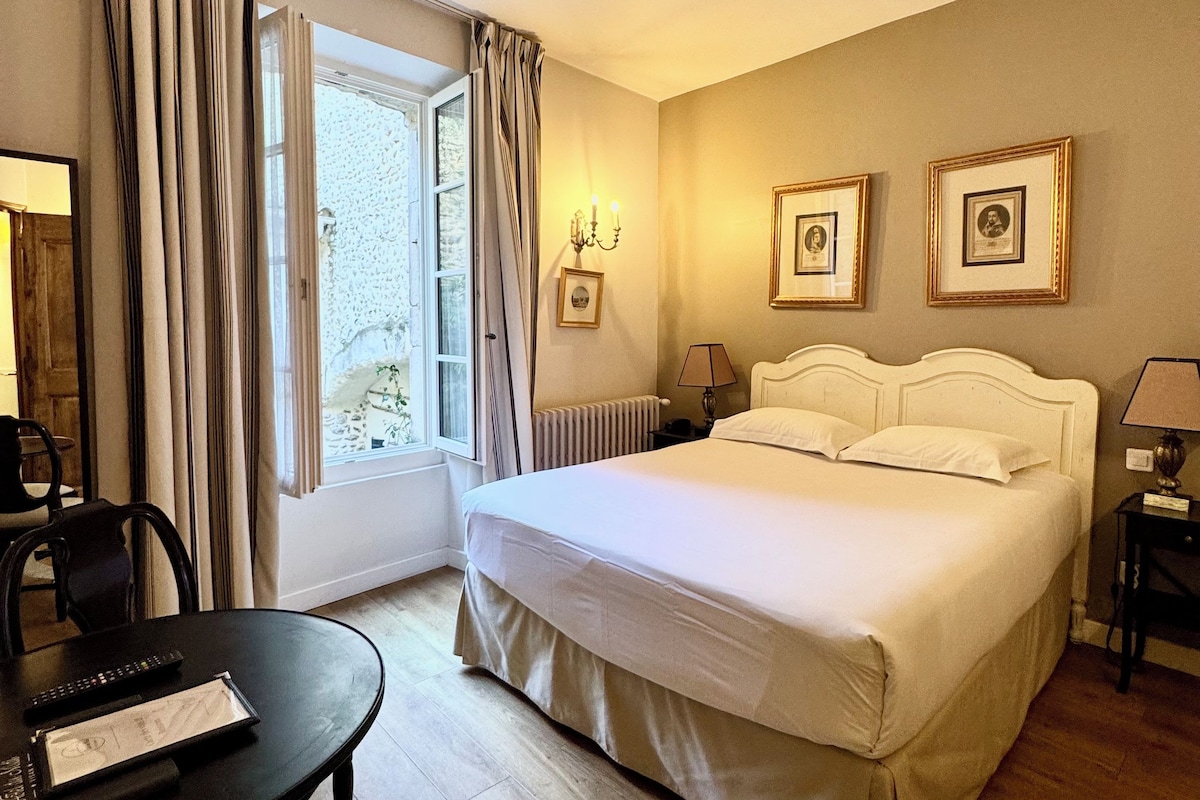A hotel room with a bed, a desk with a chair, a window with curtains, and framed pictures on the wall.