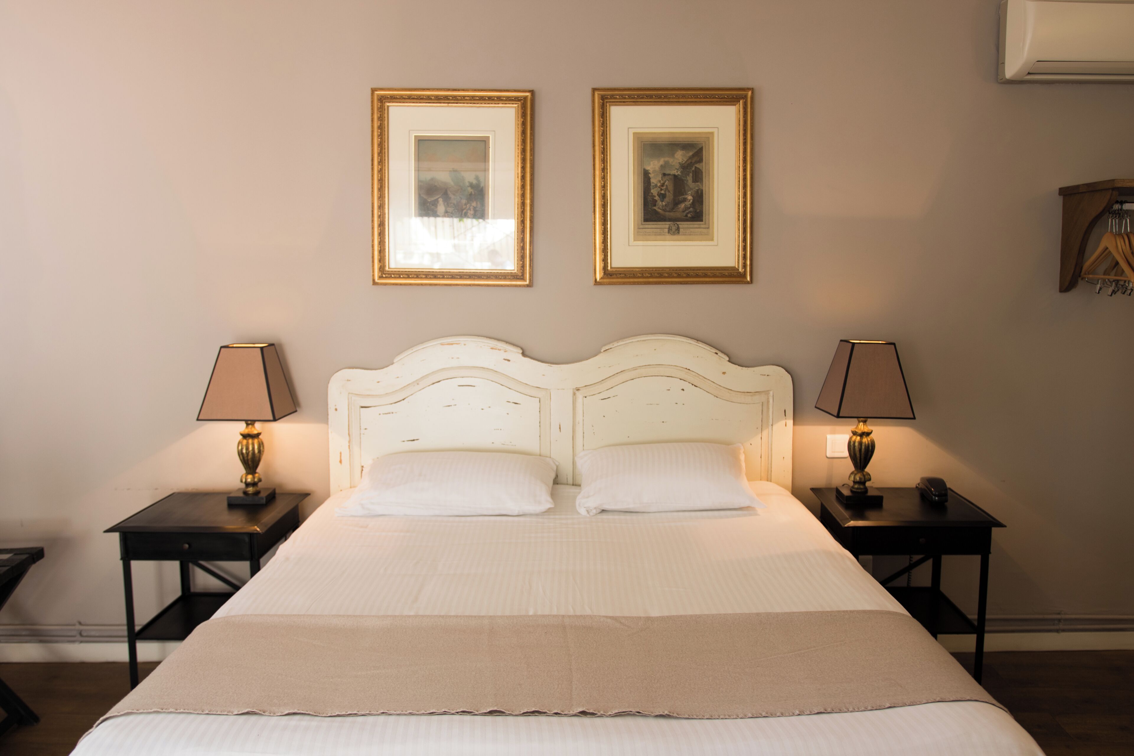 Una cama bien hecha con dos almohadas, flanqueada por dos mesitas de noche con lámparas, y dos cuadros enmarcados en la pared.