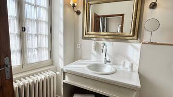Un baño con un lavamanos blanco, tocador y un espejo grande. Hay dos luces empotradas en la pared, un radiador y una ventana con cortinas.