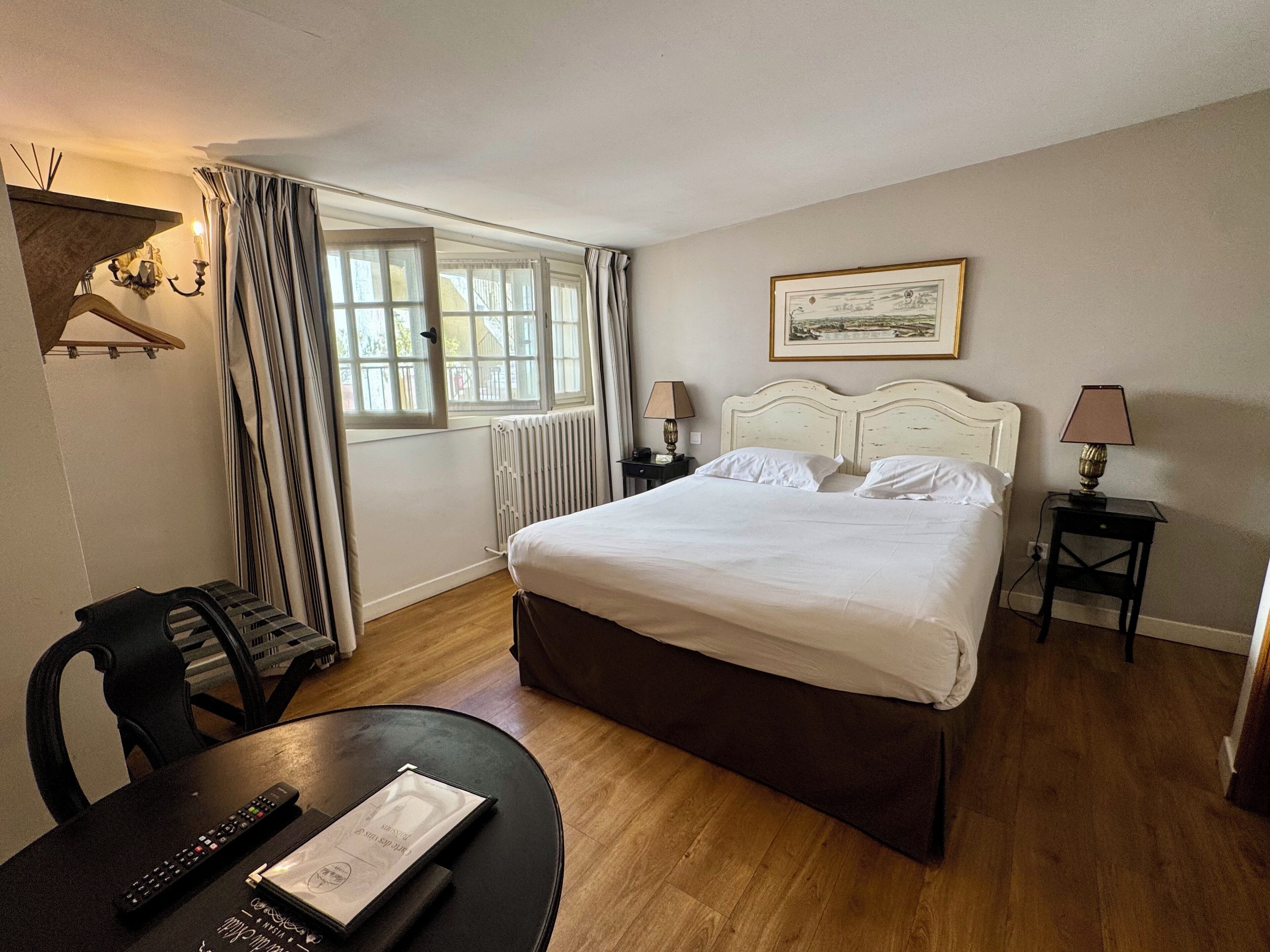 Une chambre d’hôtel avec un lit, une table avec une télécommande, une chaise, une lampe et une fenêtre avec des rideaux.