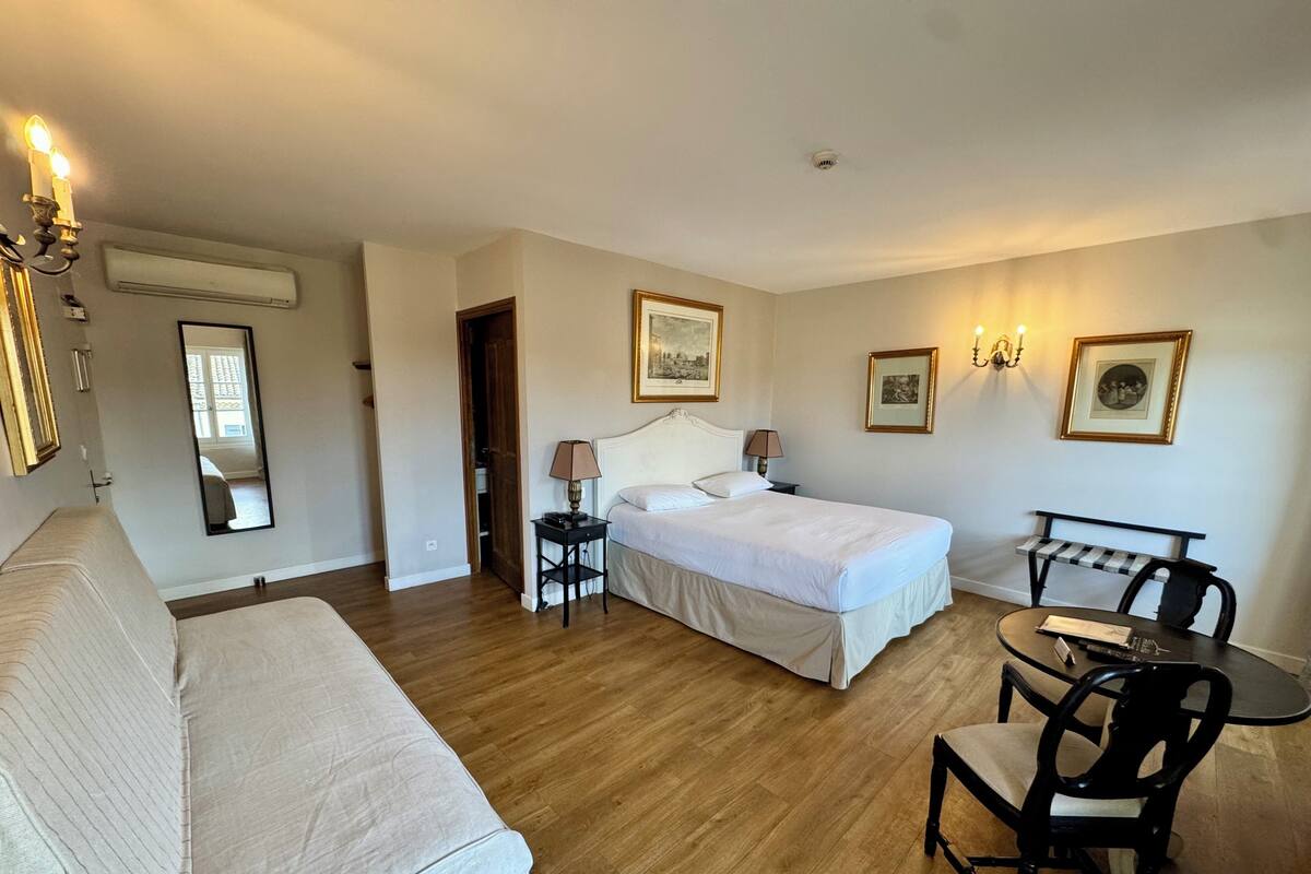 A hotel room with a bed, a sofa, a small table, and a chair. There are wall-mounted lamps and framed pictures.