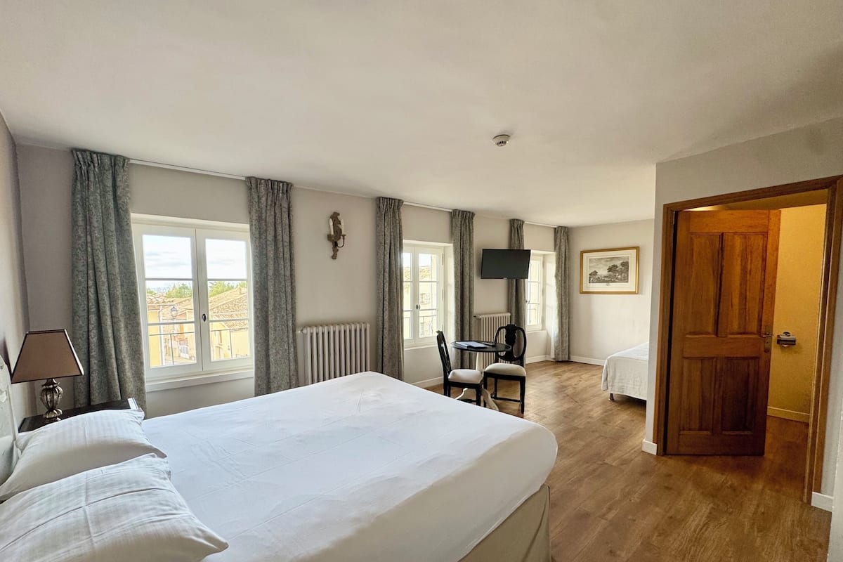 A hotel room with a large bed, a desk with a chair, a television, and a door leading to another room.