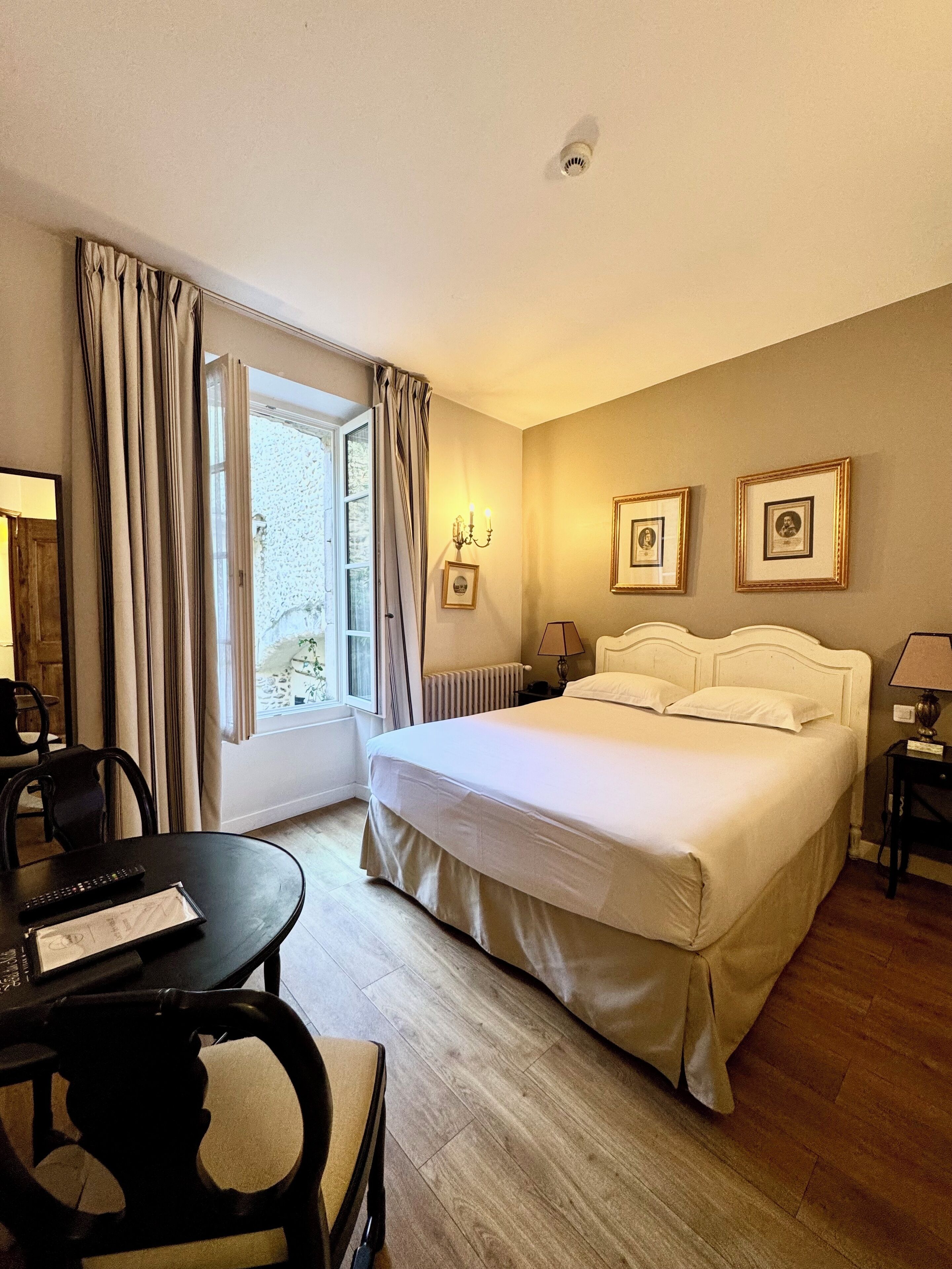 A hotel room with a bed, a desk with a chair, a window with curtains, and framed pictures on the wall.