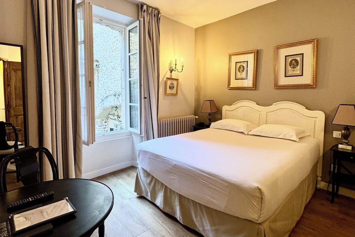 A hotel room with a bed, a desk with a chair, a window with curtains, and framed pictures on the wall.
