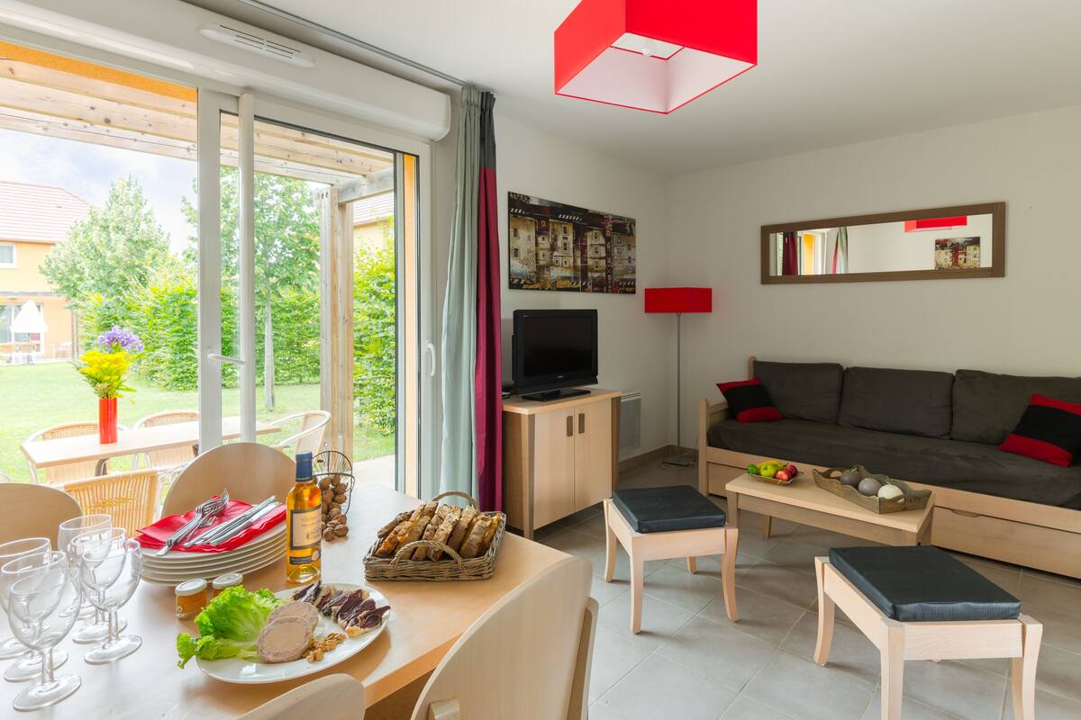 A modern living room with a sofa, coffee table, and dining area. There is a television and a window with curtains.