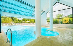 Piscina interna com tetos de vidro transparente, colunas brancas e vista para o verde lá fora.