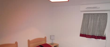A room with two single beds, a bedside table with a lamp, a window with curtains, and a wall-mounted air conditioning unit.