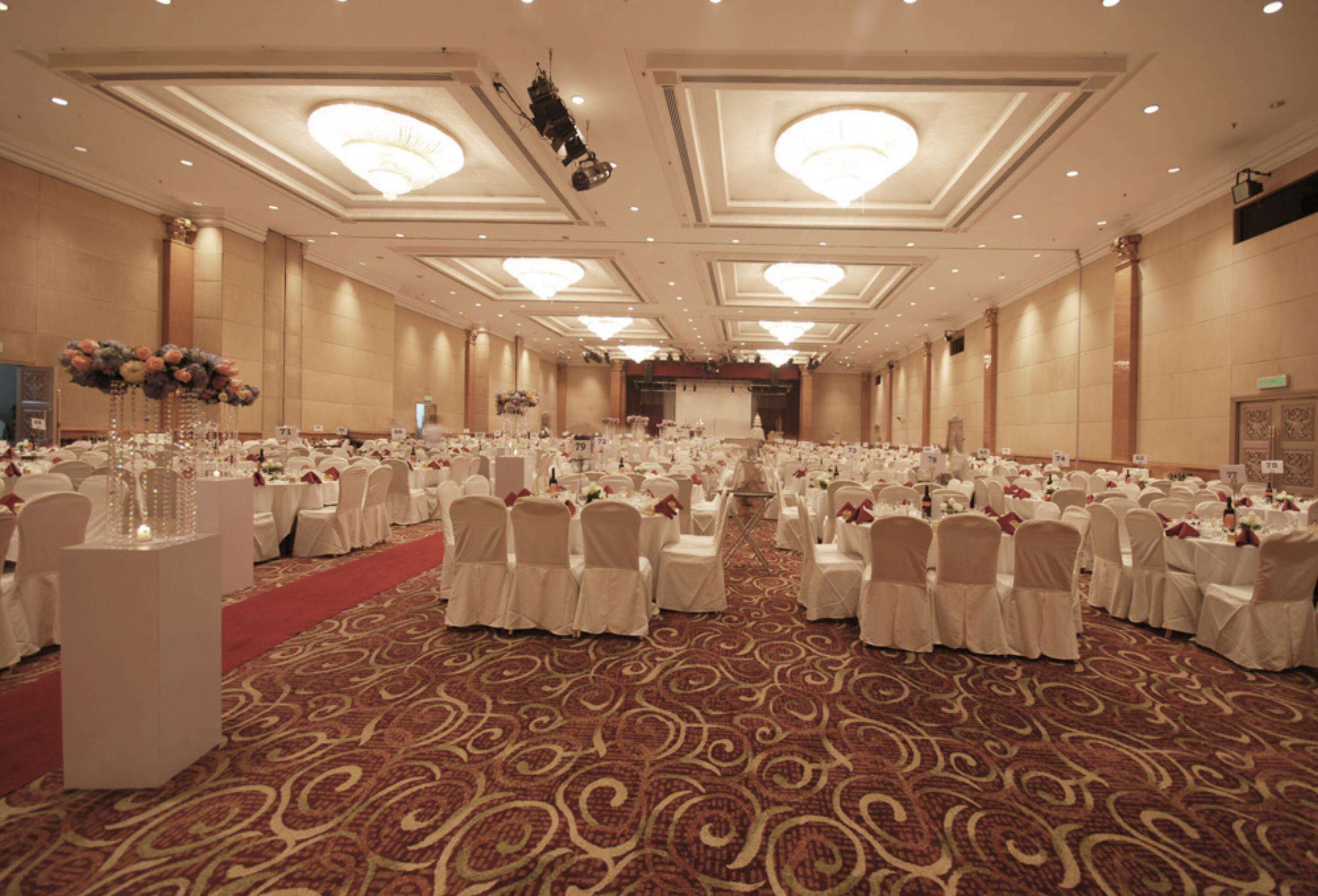 A large banquet hall with rows of round tables, white chairs, and a red carpet.