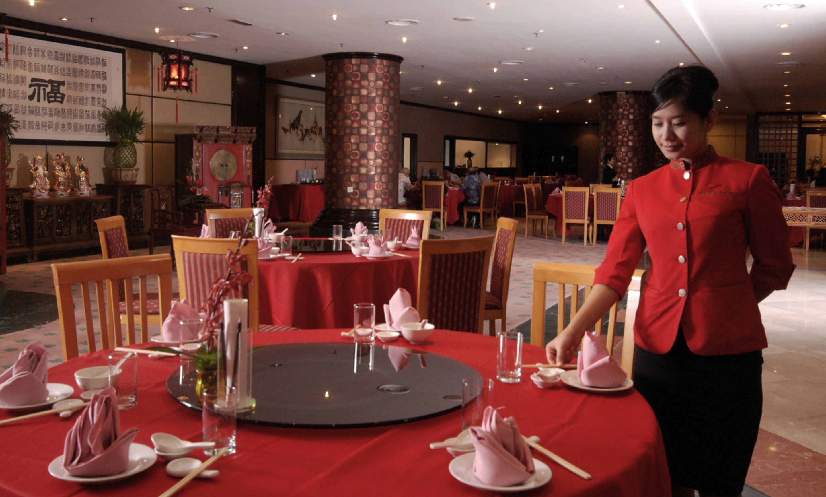 A restaurant with round tables set for dining, featuring red tablecloths and pink napkins.