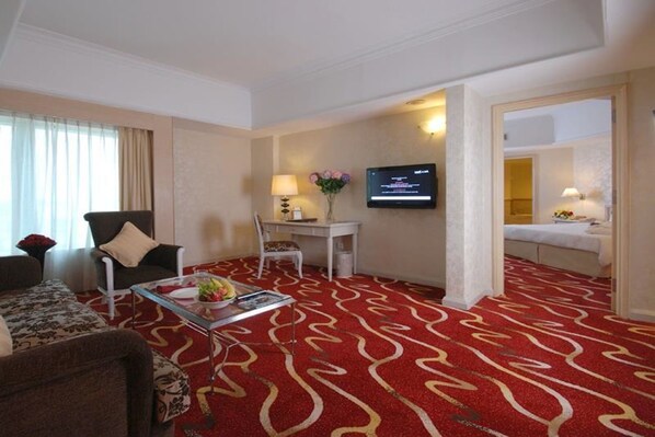 A hotel room with a red patterned carpet, a television mounted on the wall, a desk with a lamp, and a bed in the adjacent room.