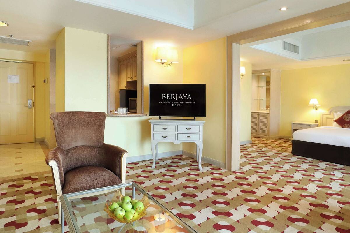 A hotel room with a bed, a TV, a chair, a small table with apples, and a patterned carpet.
