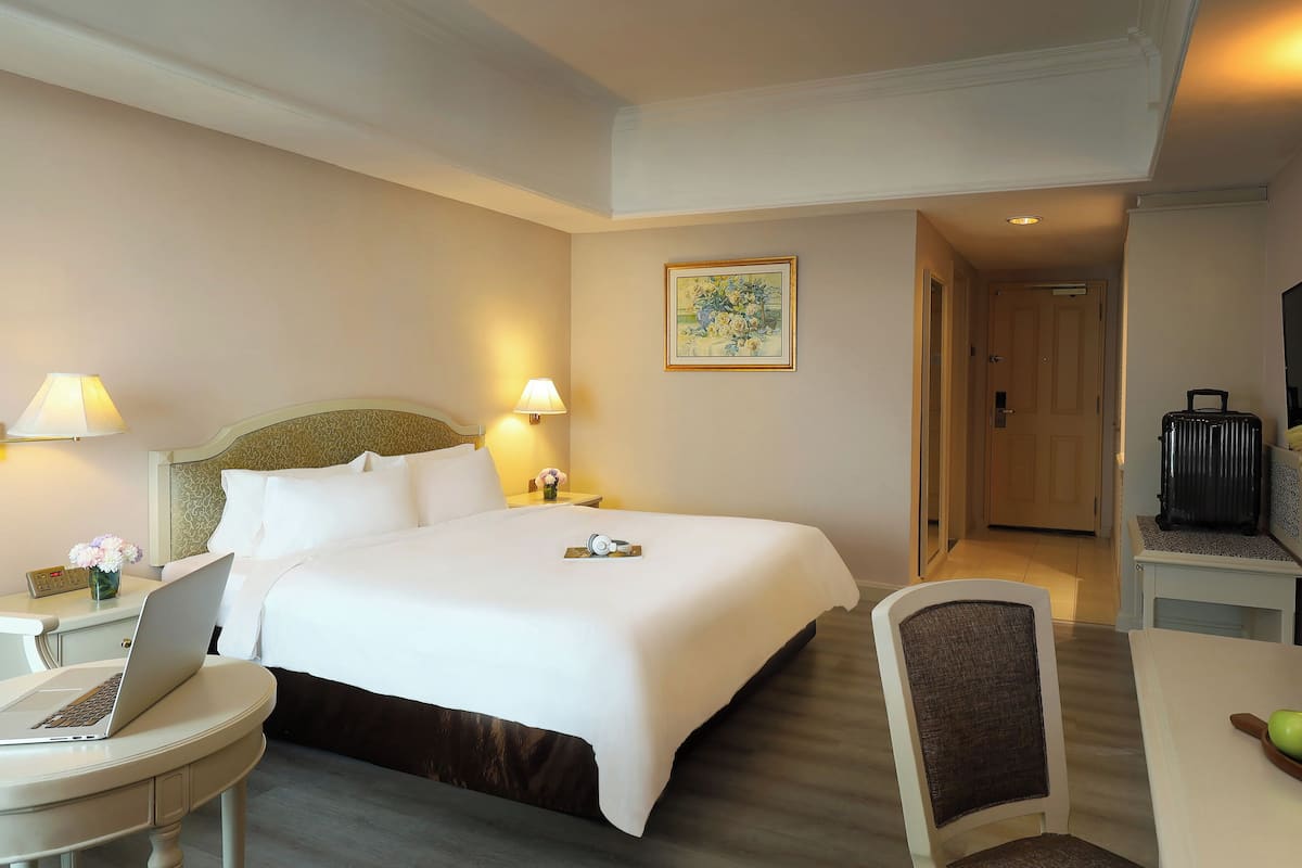 A hotel room with a large bed, bedside lamps, a laptop on a small table, a chair, and a suitcase.
