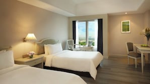 Quarto de hotel com duas camas, uma escrivaninha, uma cadeira, um abajur e uma janela com vista para a cidade.