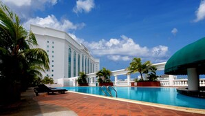 Piscina com espreguiçadeiras e palmeiras, cercada por um edifício branco sob um céu azul.