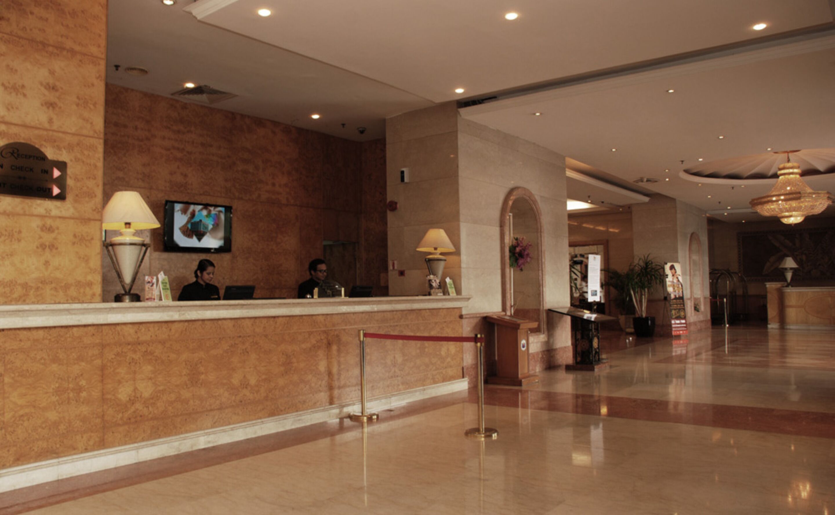 A reception area with a marble counter, two receptionists, a television screen, and a chandelier.