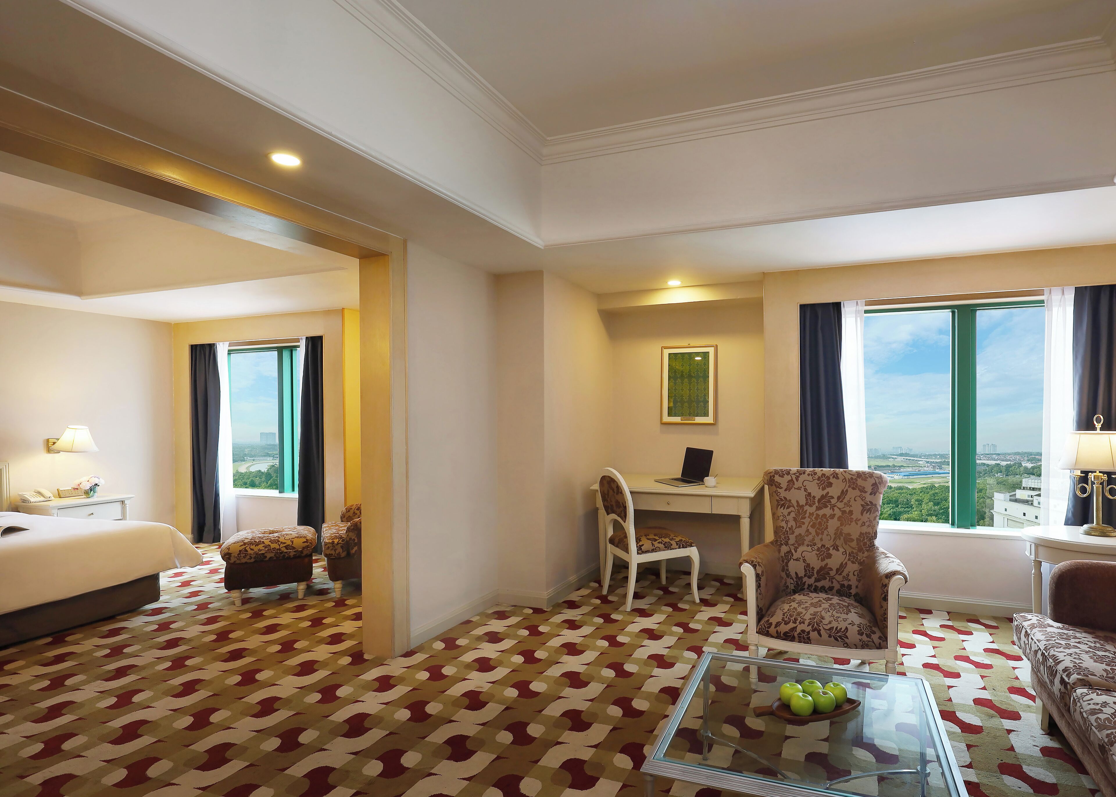 A hotel room with a bed, a desk with a laptop, a chair, a sofa, a coffee table with apples, and a view of the city through large windows.