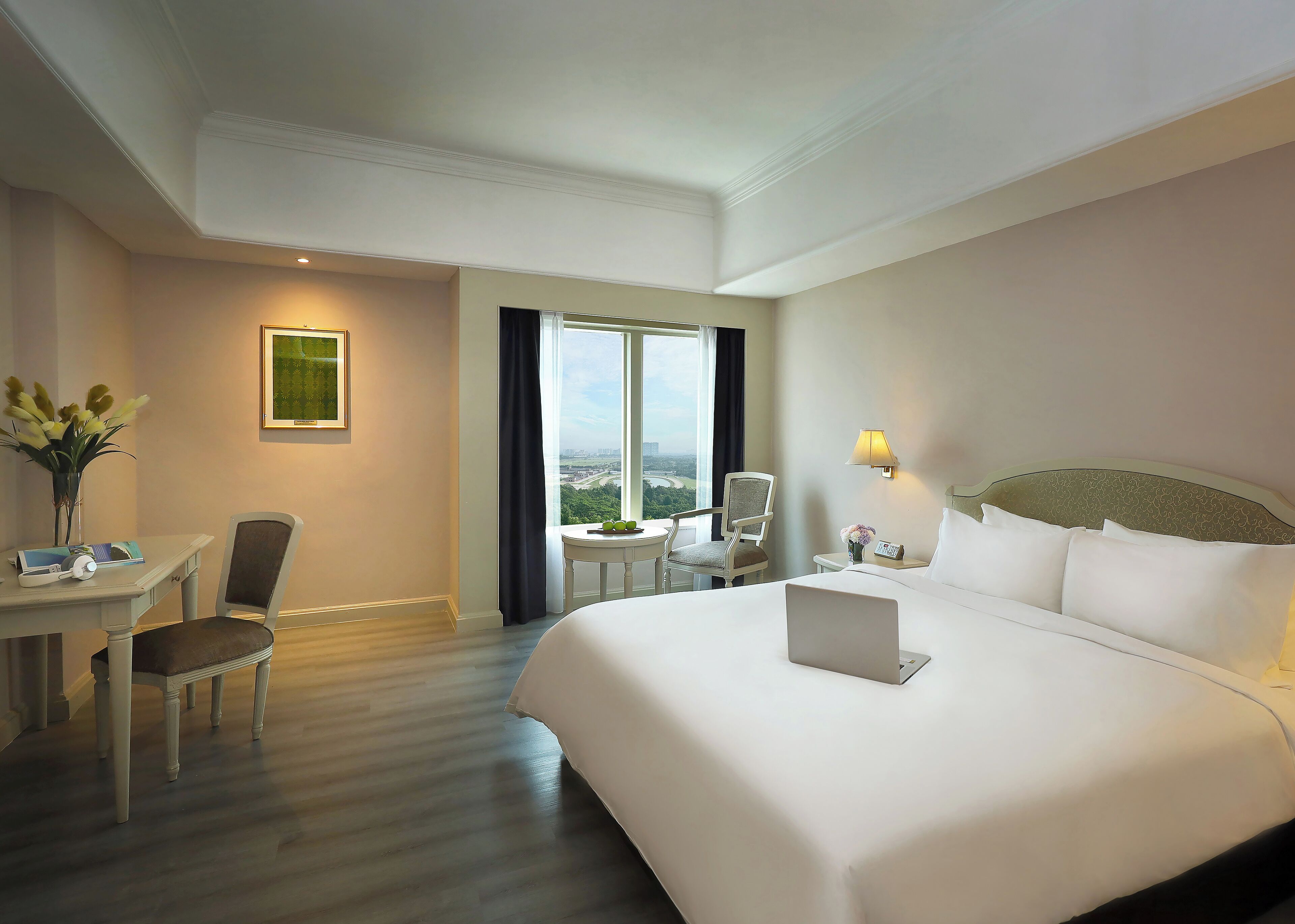 A hotel room with a large bed, a desk with a chair, a small table with a lamp, and a view of the city through the window.