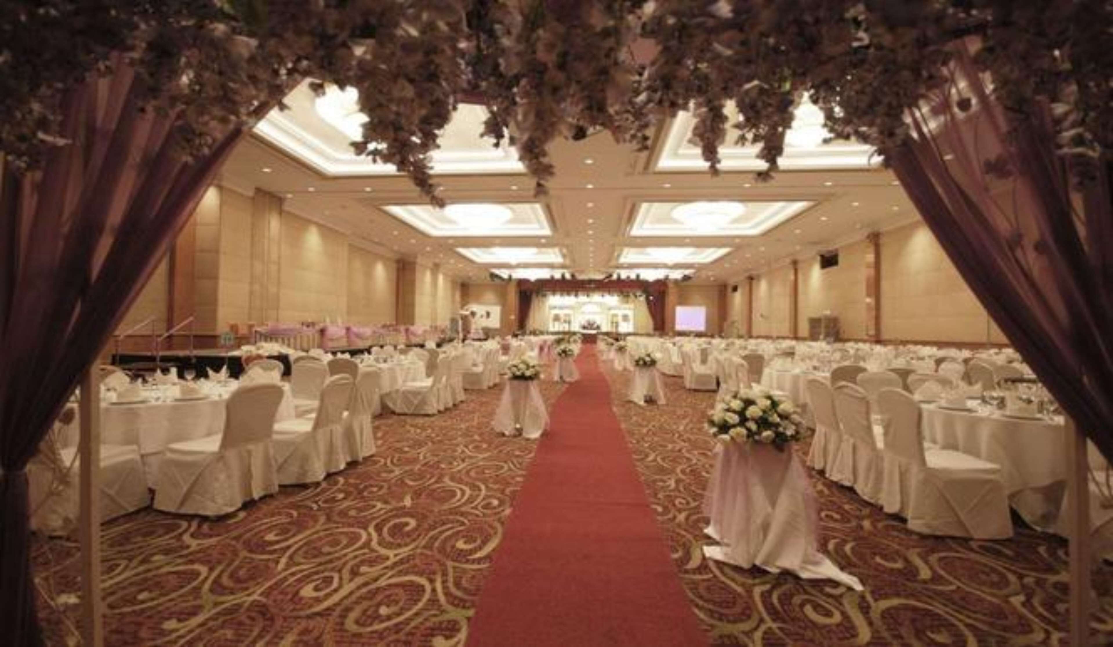 Un amplio salón de banquetes con hileras de mesas redondas, manteles blancos y sillas. Una alfombra roja recorre el centro.