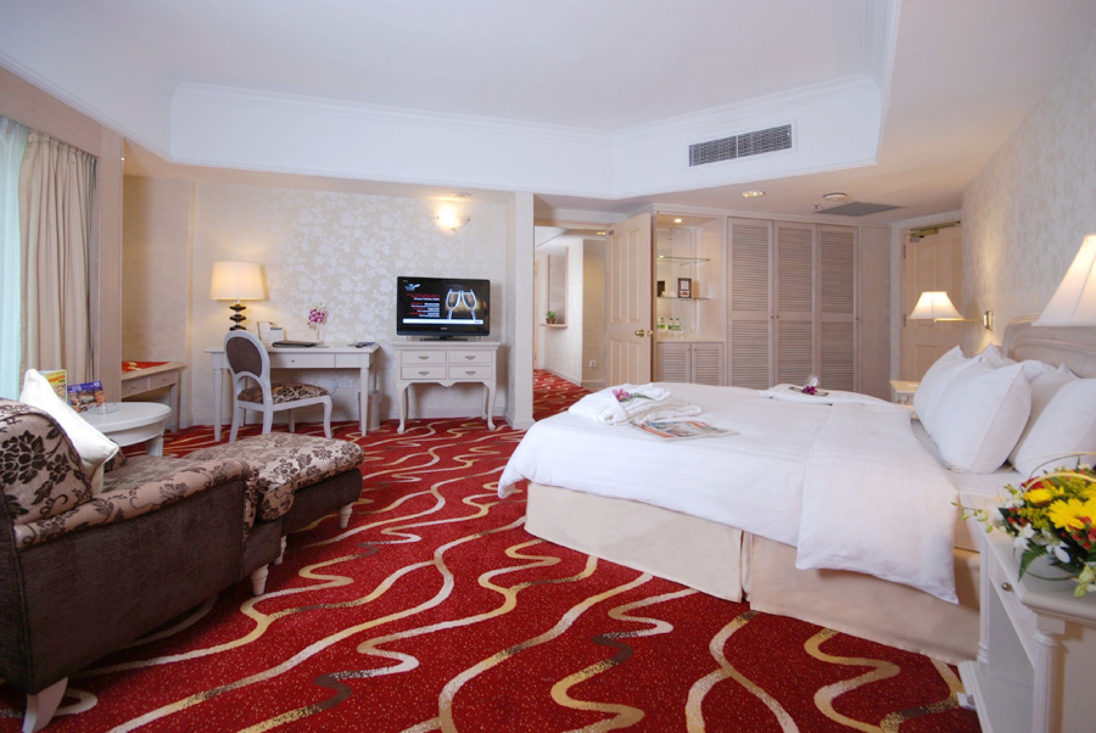 A hotel room with a large bed, a desk with a TV, a chair, and a red carpet.