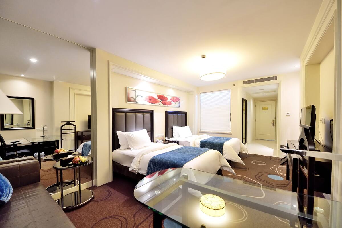 A hotel room with two beds, a TV, a glass table, a sofa, and a desk with a lamp.