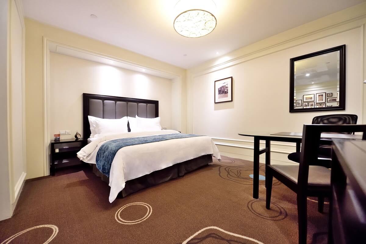 A hotel room with a bed, bedside table, chair, and a desk with a chair.